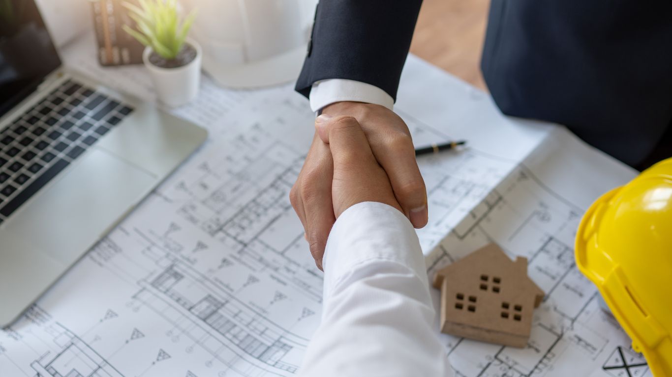 Two hands shaking with a yellow protective hat and a house plan with a laptop beside.